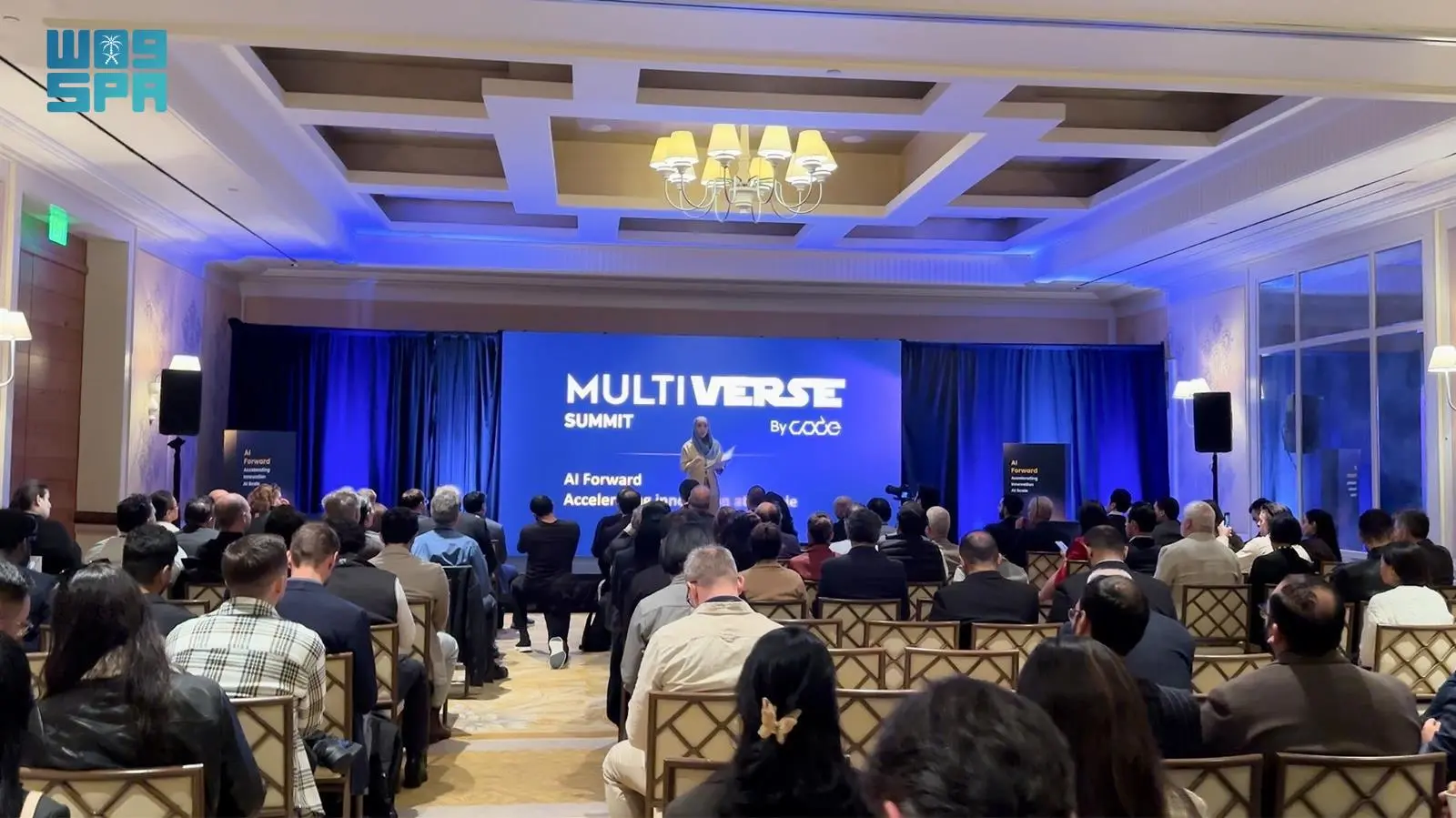 A speaker on stage addressing a large audience seated in rows at the Multiverse Summit with a large screen displaying "MULTIVERSE SUMMIT by Code" in a conference room with blue lighting.