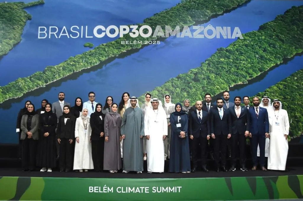 A large group of people, including men in traditional white thobes and women in abayas and hijabs, as well as others in suits and business attire, pose for a group photo on a stage. Behind them is a large screen displaying 'BRASIL COP30 AMAZÔNIA BELÉM 2025' with an aerial view of a river winding through a lush green forest. The stage also has 'BELÉM CLIMATE SUMMIT' written at its base.
