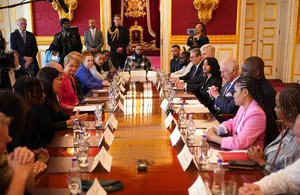 Idris Elba, Prime Minister Rishi Sunak, and other government officials and young people sit around a large, polished wooden table in an ornate room, engaged in a discussion. Elba and Sunak are seated opposite each other near the center of the table. The room features high ceilings, decorative gold accents, and large paintings, with several other individuals standing in the background.