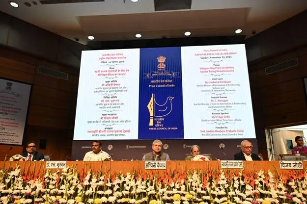 Union Minister Ashwini Vaishnaw seated on a stage with other dignitaries, including Dr. L. Murugan, during the National Press Day 2025 celebration in New Delhi, with a large screen displaying event details in the background.