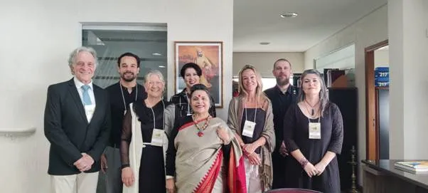 A diverse group of eight individuals, four men and four women, stand together and smile in an indoor setting with a framed picture on the wall behind them.