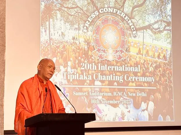 A Buddhist monk in orange robes speaks at a podium during a press conference for the 20th International Tipitaka Chanting Ceremony. Behind him, a screen displays the event's logo and details, including '20th International Tipitaka Chanting Ceremony' and 'Samvet Auditorium, IGNCA, New Delhi, 16th November, 2025'.