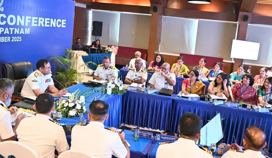 A group of Indian Navy officers and educators, including men and women, are seated around a large blue-draped conference table in a well-lit room. A sign in the background reads 'CONFERENCE VISAKHAPATNAM NOVEMBER 2025'. Several officers in white uniforms are at the head of the table, while other participants are on the sides, some taking notes.