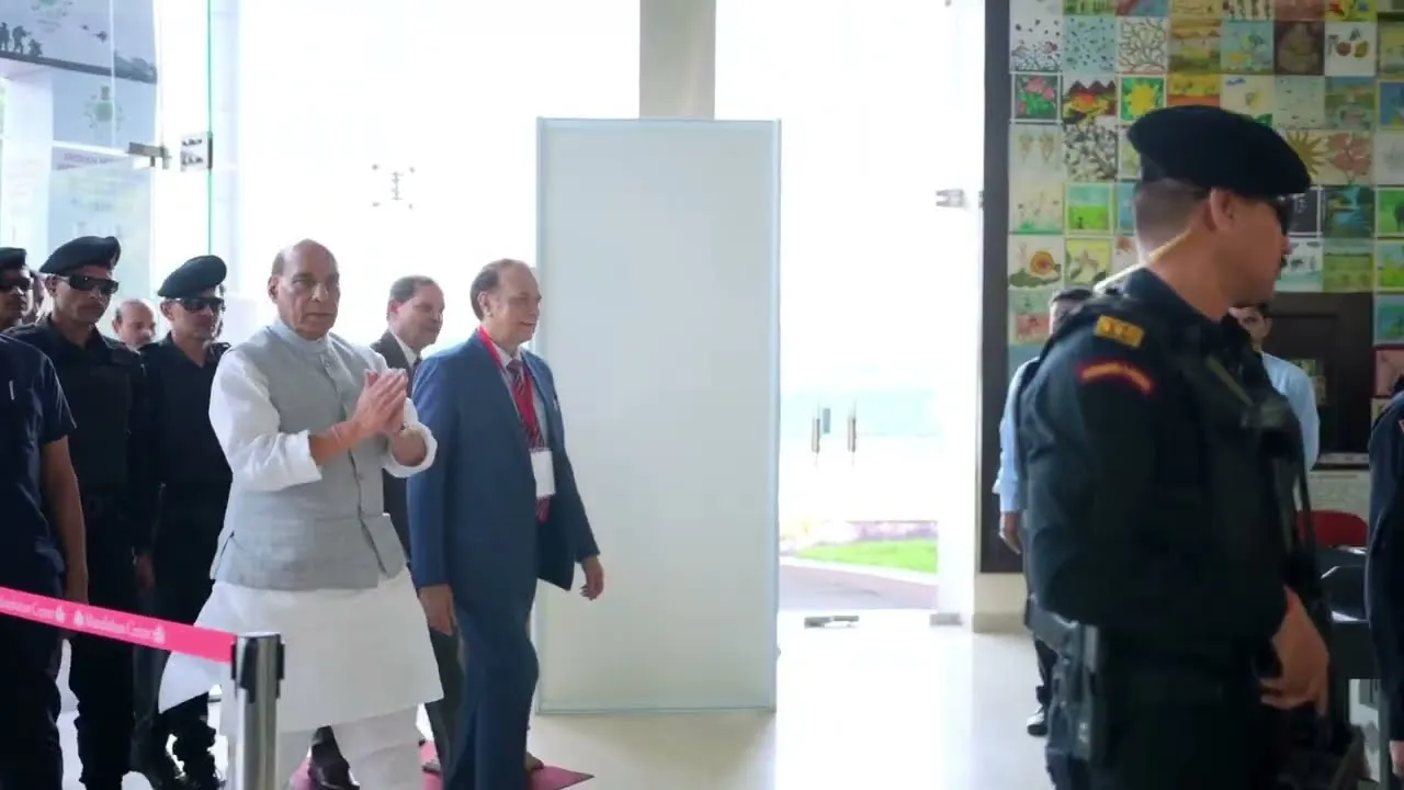 India's Defence Minister Rajnath Singh, dressed in a white kurta and grey waistcoat, claps while walking through a brightly lit indoor space with another man in a blue suit beside him. Security personnel in black uniforms and berets are visible around them, with a wall featuring children's drawings in the background.