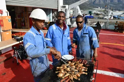 Three male fishermen in blue overalls and white hard hats are grilling meat on a portable barbecue on the red deck of a fishing vessel.