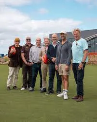 Seven men, including Australian Prime Minister Anthony Albanese and Victorian Premier Jacinta Allan, stand on a green sports field. Albanese holds a football.