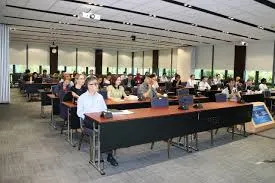 A wide shot of a brightly lit conference room filled with diverse attendees seated at long tables, facing forward. Some individuals are looking towards the front where a screen or speaker would be, while others are engaging with materials on their tables. The room has large windows with blinds and a projector visible on the ceiling.