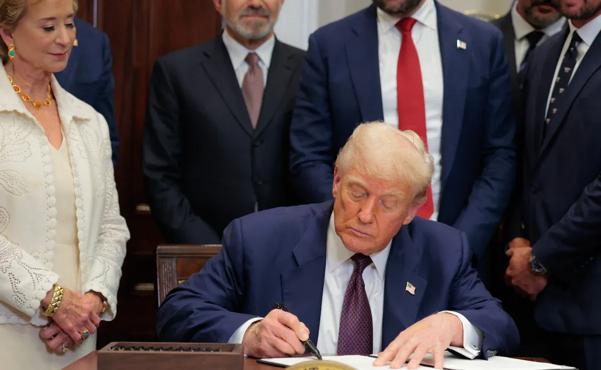 Donald Trump signing documents at a desk with several people observing him, including a woman in a white jacket to his left.