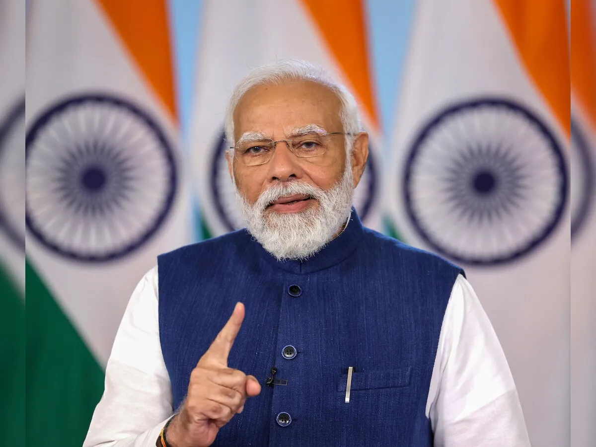 Indian Prime Minister Narendra Modi, a man with a white beard and spectacles, wearing a white kurta and a blue sleeveless jacket, points his right index finger forward while speaking. He is standing in front of two blurred Indian flags.