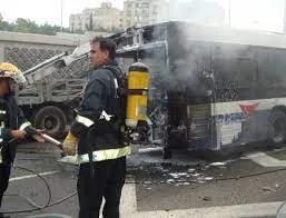 A firefighter in protective gear with an oxygen tank on his back stands next to a smoking, burnt-out bus, holding a hose. Another person in fire protective clothing is partially visible on the left. The scene appears to be an urban or roadside environment with buildings in the background.