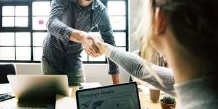 Two people, one in a patterned button-up shirt and another with blonde hair, shaking hands over a table with a laptop and documents in an office setting.