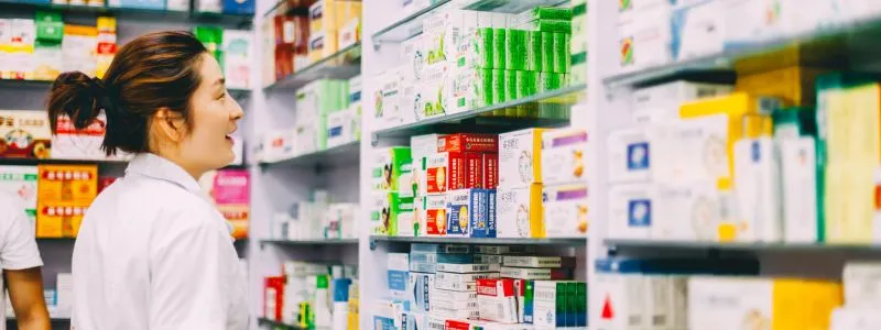 A female nurse practitioner in a white lab coat looks at shelves stocked with various packaged medicines and health products in a pharmacy setting.