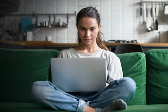 A young woman with dark hair, wearing a light grey t-shirt and blue jeans, sits cross-legged on a dark green sofa. She is looking intently at a silver laptop open on her lap, with her hands resting on the keyboard. The background shows a modern kitchen with white tiled walls and various kitchen utensils.