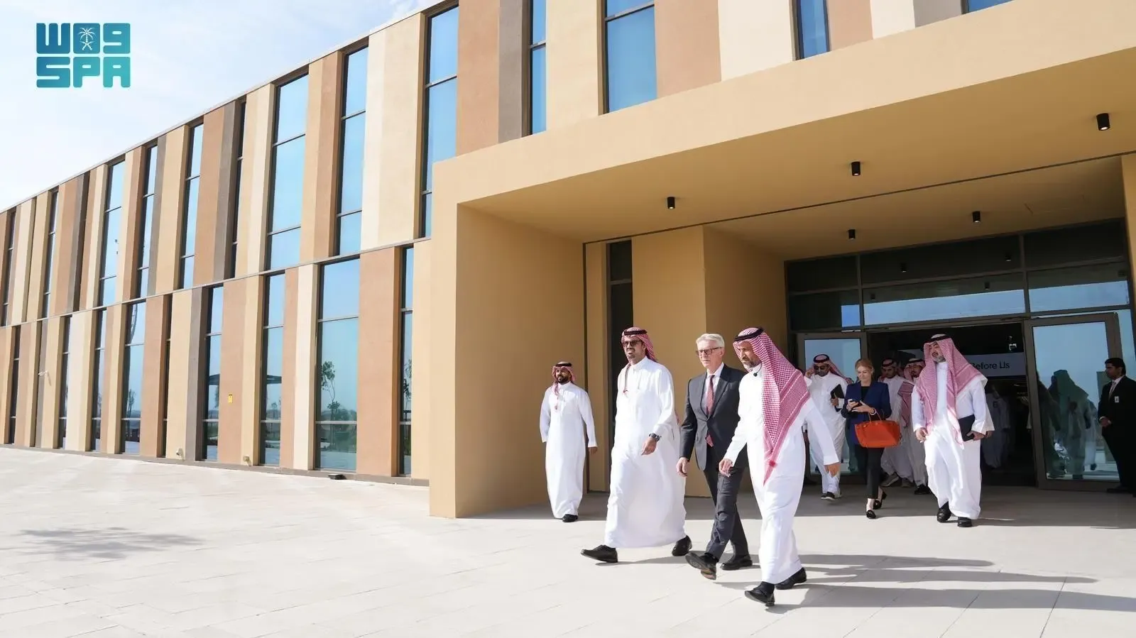 Several men in suits and traditional Saudi attire walk outside a modern, multi-story building. The building has a tan facade with large rectangular windows and a prominent entrance. The sky is clear and blue.
