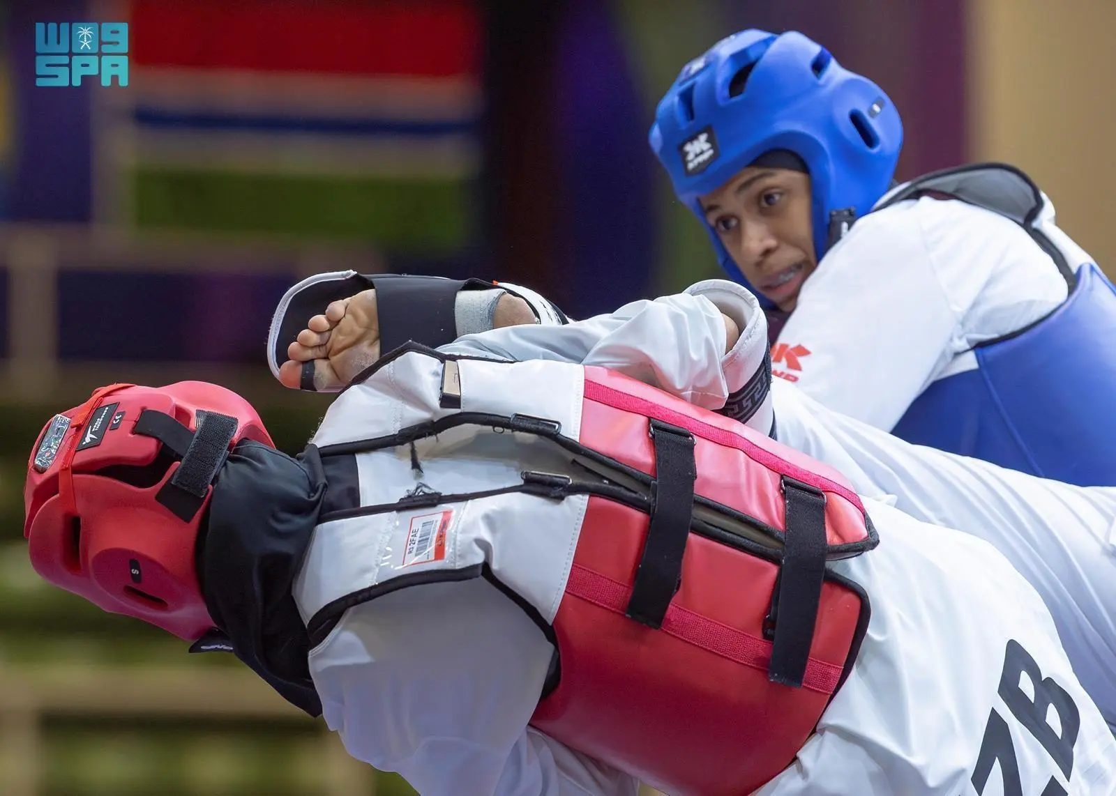 A Saudi female taekwondo athlete in a blue helmet and white uniform with blue padding kicks her opponent, who is wearing a red helmet and red chest protector, during a match. The kicking athlete's foot is visible near the opponent's chest protector, indicating a direct hit or an attempted strike.