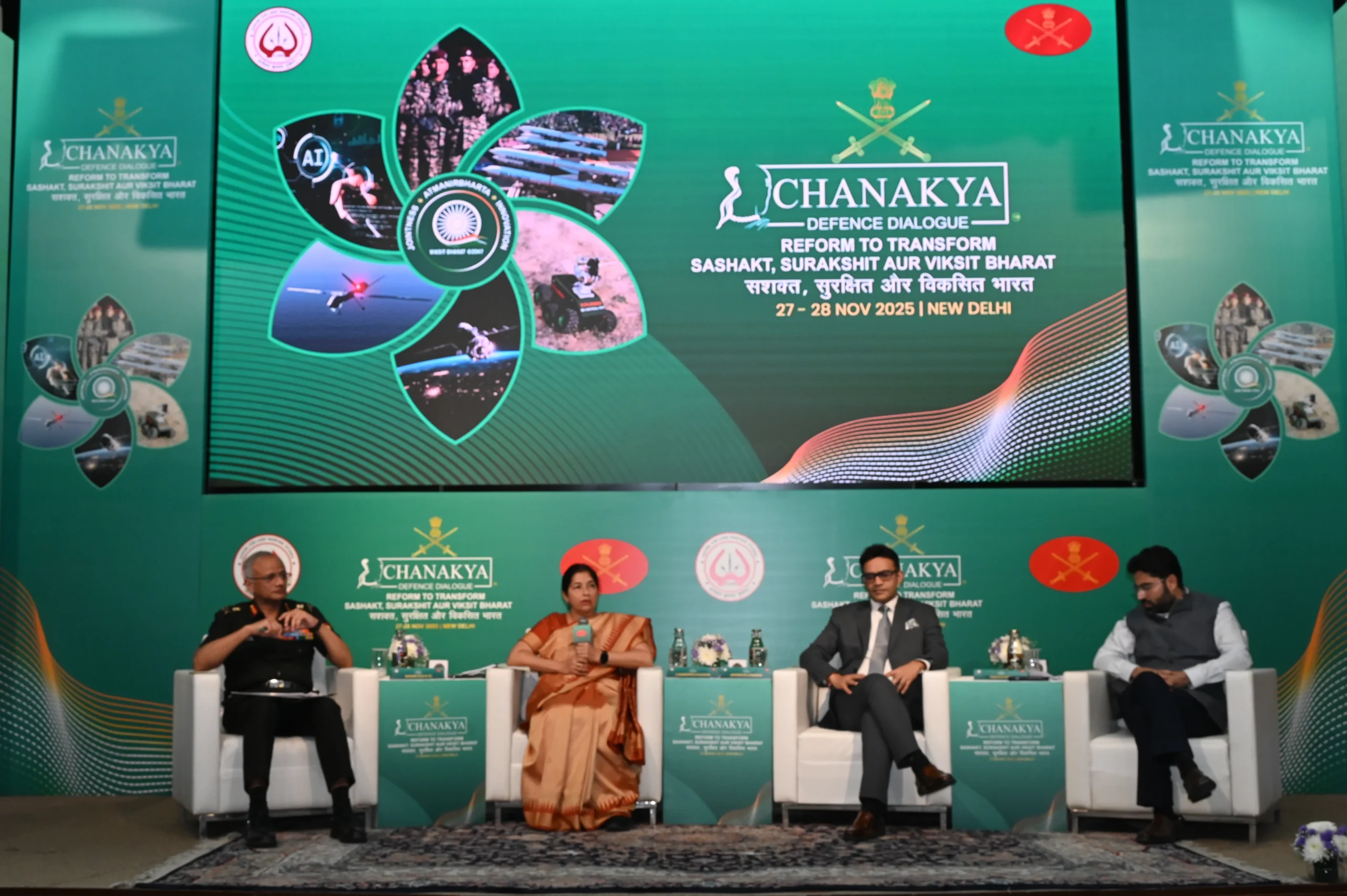 Four panelists, three men and one woman, sit on white armchairs on a stage with a large green digital backdrop displaying the "Chanakya Defence Dialogue 2025" logo and event details. The panelists are formally dressed, and the stage is set for a discussion.