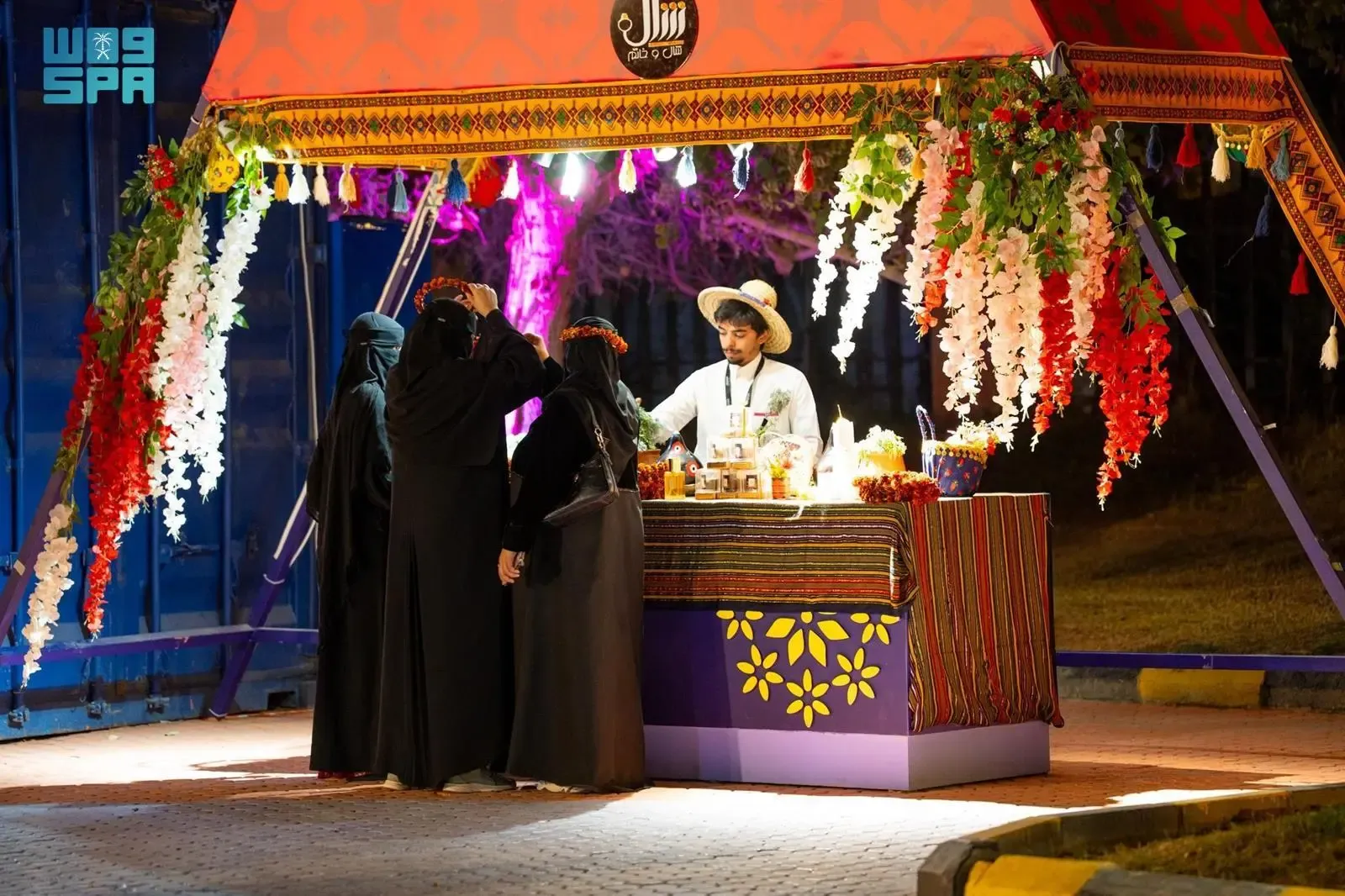 A man in a straw hat is serving drinks from a colorful, decorated stall to several women wearing abayas and flower crowns at an outdoor market event at night. The stall is adorned with flowing red and white flowers and tassels, and the man is handing a drink to one of the women. The overall setting is vibrant and festive, with warm lighting.