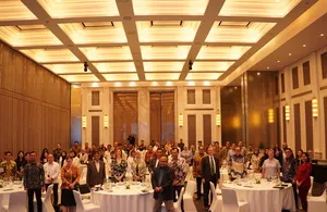 A large group of people stand in a brightly lit conference room with tables set for a meal, attending the RIA Forum 2025 in Jakarta.