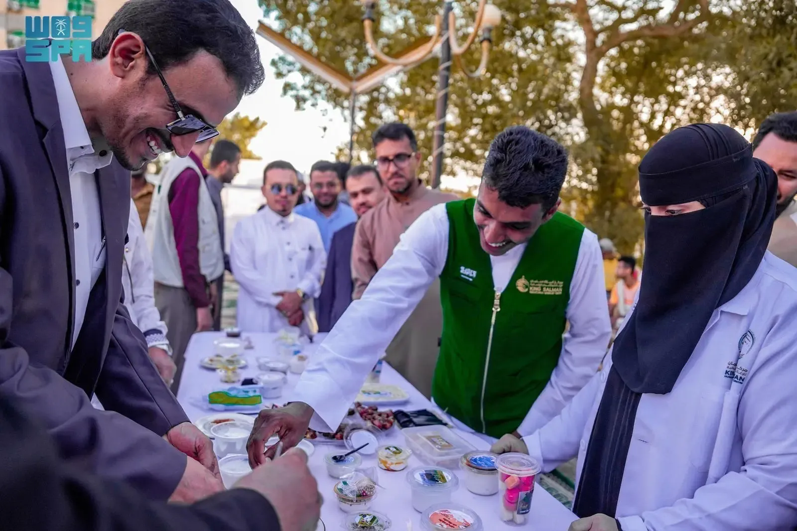 A group of people, including men in traditional Saudi attire and a woman in a black niqab and white lab coat, are gathered around a table with small food samples and products. A man in a green vest with a logo is interacting with others, while another man in a suit smiles. The setting appears to be an outdoor market or exhibition with trees and other people in the background.