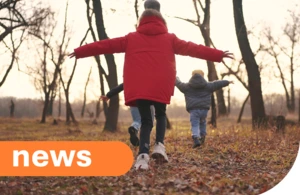 Three children, seen from behind, walk through an autumn forest. The child in the foreground wears a red winter coat and has their arms outstretched for balance. The ground is covered with fallen leaves and dry grass, and bare trees are visible in the background under a warm, soft light.
