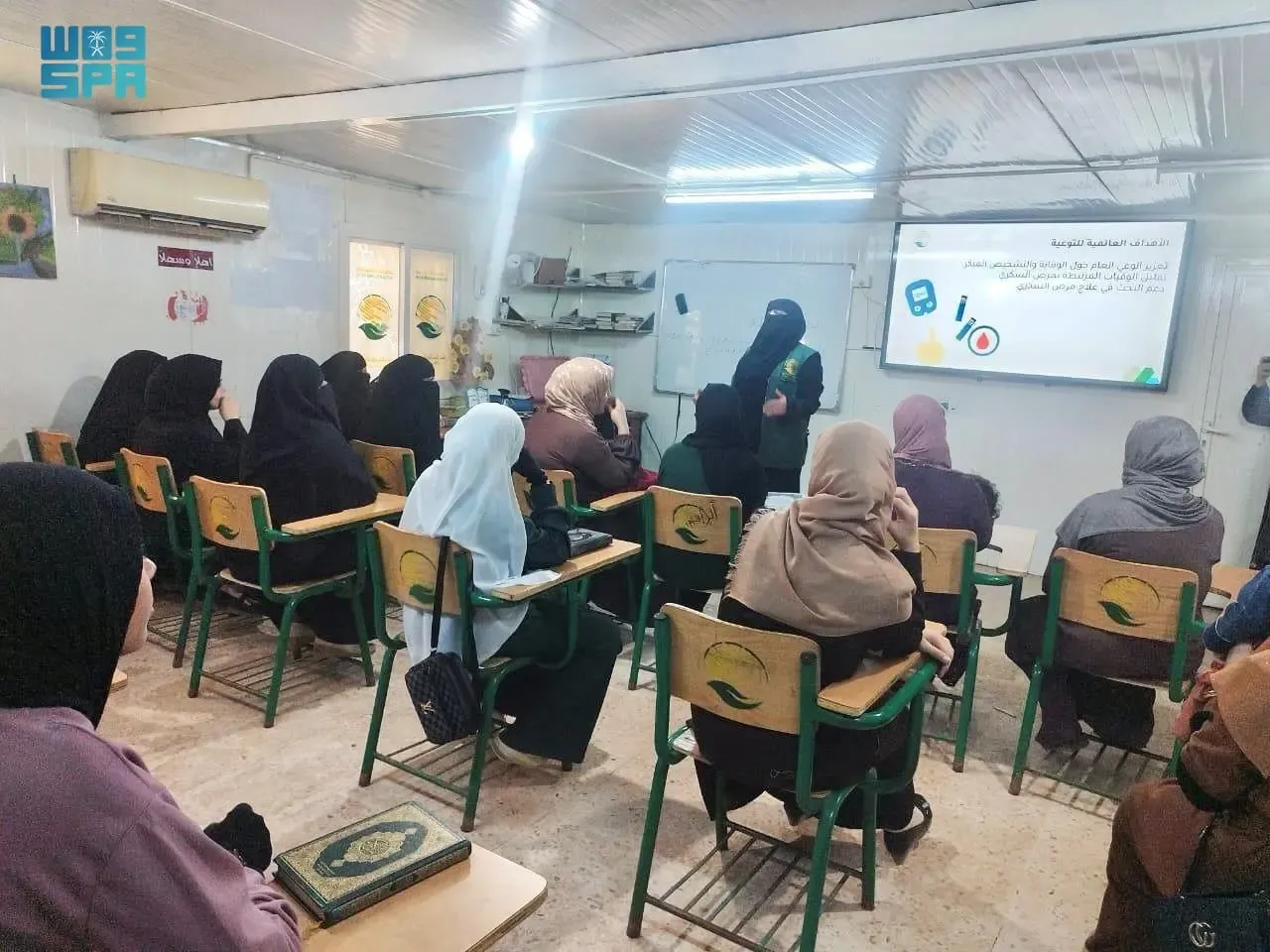 A classroom scene where several women, most wearing headscarves, are seated at desks, facing a projector screen at the front. A woman in a dark abaya and niqab is standing near the screen, appearing to give a presentation. The screen displays text in Arabic and a graphic related to diabetes awareness, showing a blood glucose meter and a food pyramid. The classroom is modestly furnished, with a whiteboard, a clock, and some decorations on the walls.