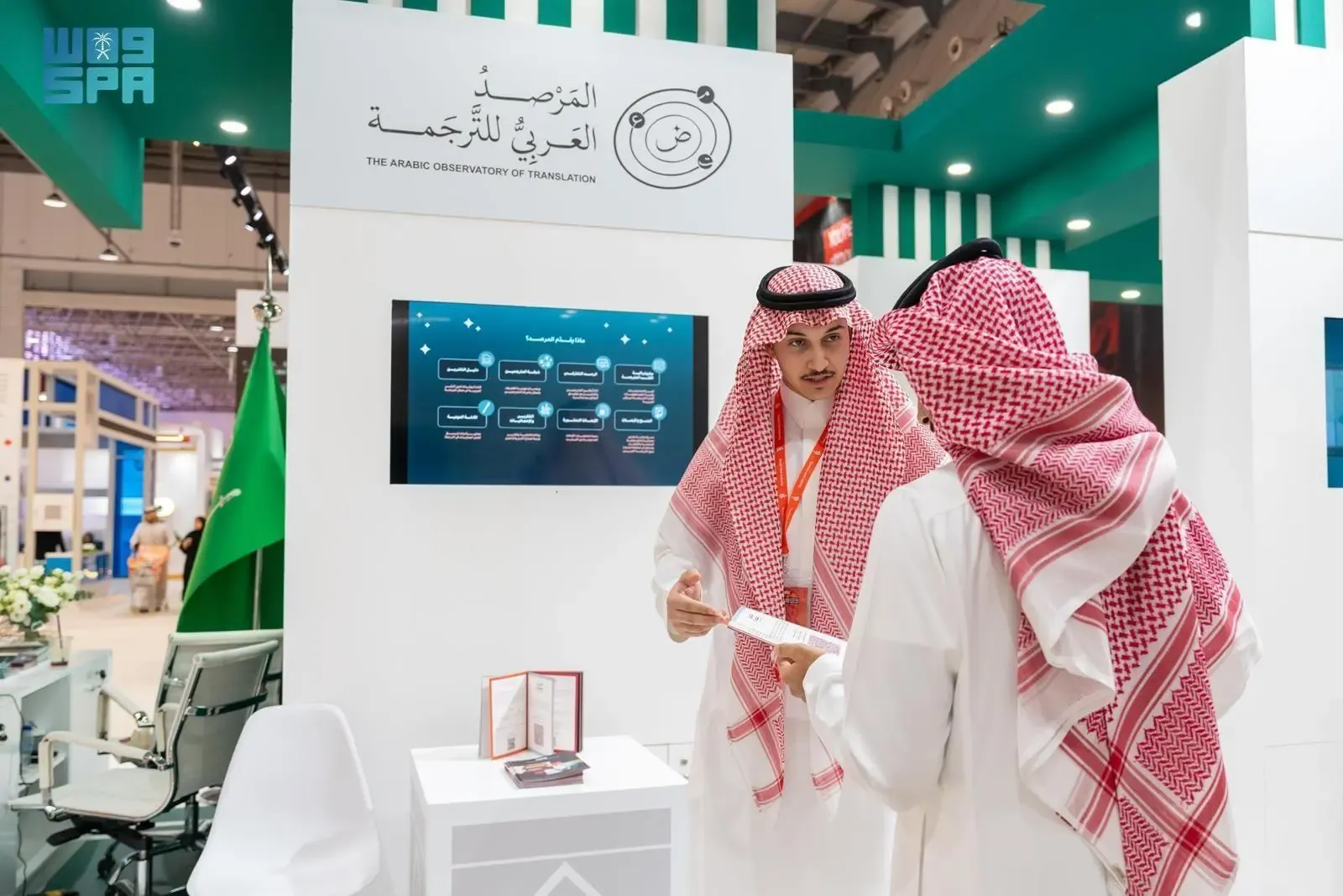 Two Saudi men in traditional thobes and ghutras standing in front of a booth for "The Arabic Observatory of Translation" at an exhibition. One man is gesturing towards a display screen.