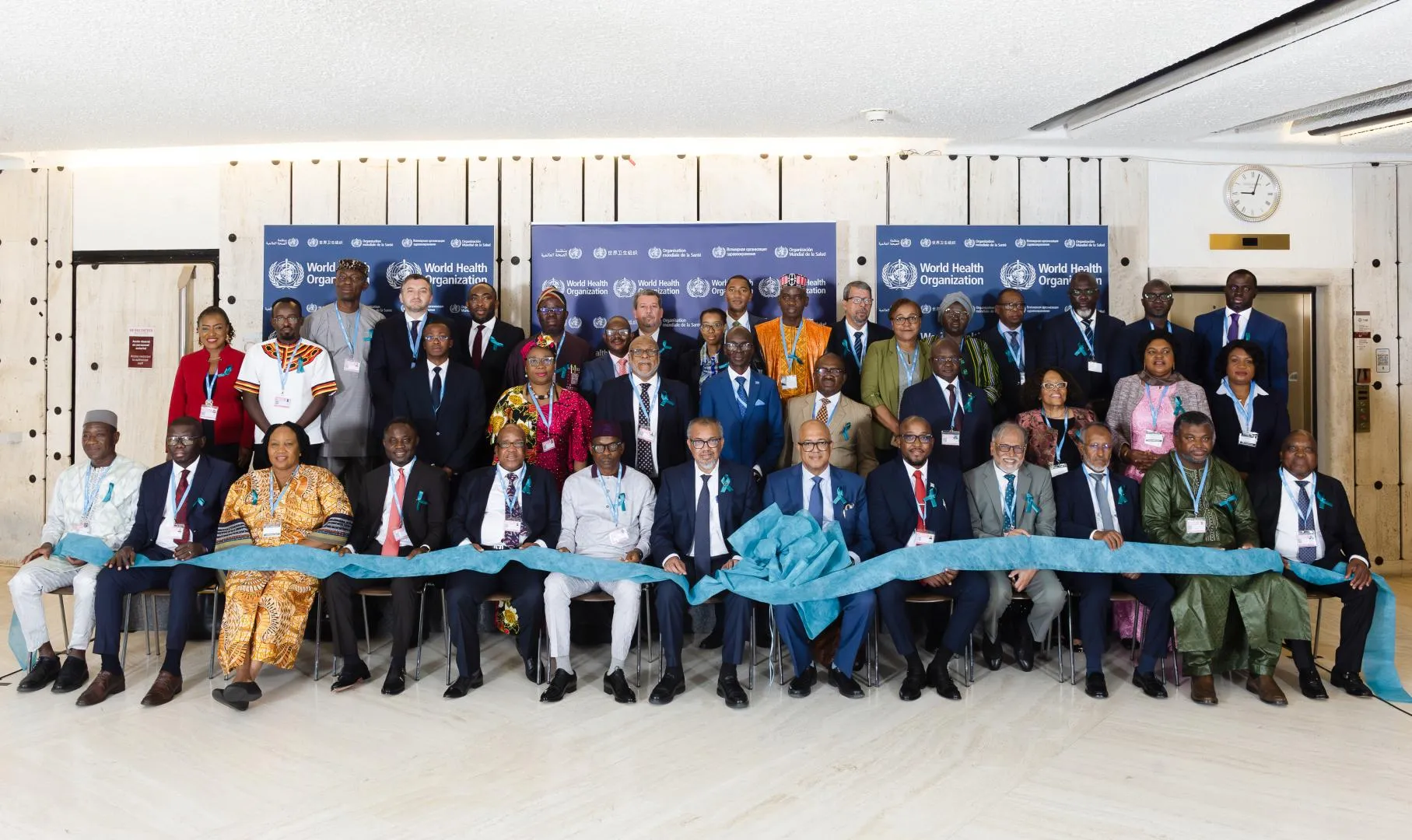 A large group of men and women, dressed in formal attire with teal ribbons pinned to their clothes, gather for a photo. They are holding a long, teal ribbon across the front, with some individuals holding scissors, suggesting a ribbon-cutting ceremony. In the background, there are light blue banners with "World Health Organization" logos.