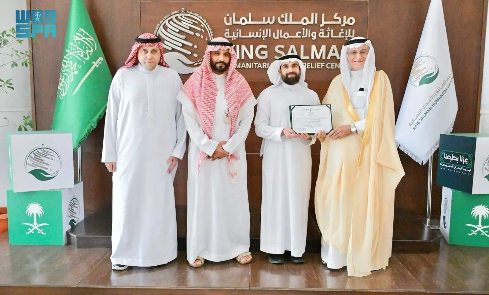 Four Saudi men in traditional thobes stand in a formal setting, with two holding a certificate, against a backdrop featuring the King Salman Humanitarian Aid and Relief Centre (KSrelief) logo and Saudi flags.