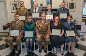 Ten individuals, some in military uniforms and some in civilian attire, stand and sit holding commendation certificates in a formal room. There are seven men and three women. The background features framed portraits and classical decor.