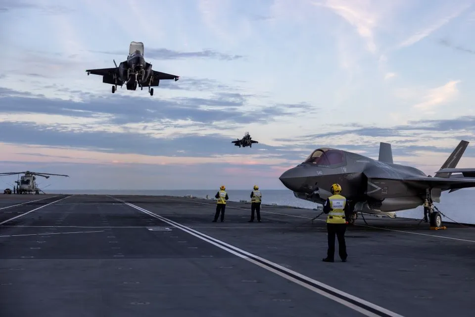 Several F-35 fighter jets are on the deck of an aircraft carrier, with two more F-35s in the air, one ascending and one descending. Naval personnel in yellow vests are visible on the deck. A helicopter is also visible in the background on the left.