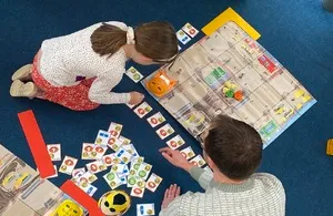 A high-angle shot shows two people, a child and an adult, playing a board game on a blue carpet. They are surrounded by numerous small cards with numbers and symbols. The child, wearing a white patterned top and red skirt, is on the left, looking down intently at the game board. The adult, in a grey long-sleeved shirt, is on the right, also focused on the game.