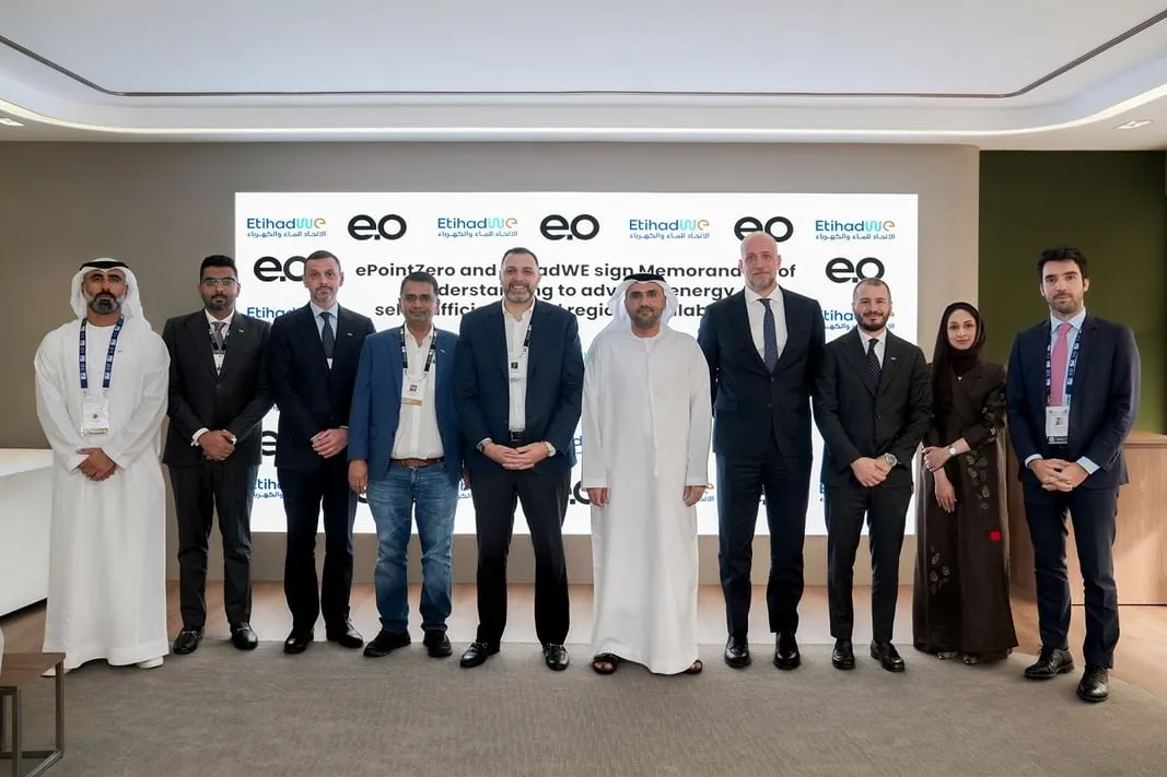 A group of ten men in business attire and traditional Emirati dress stand in a line smiling, posing for a photo in front of a white screen with company logos and text.