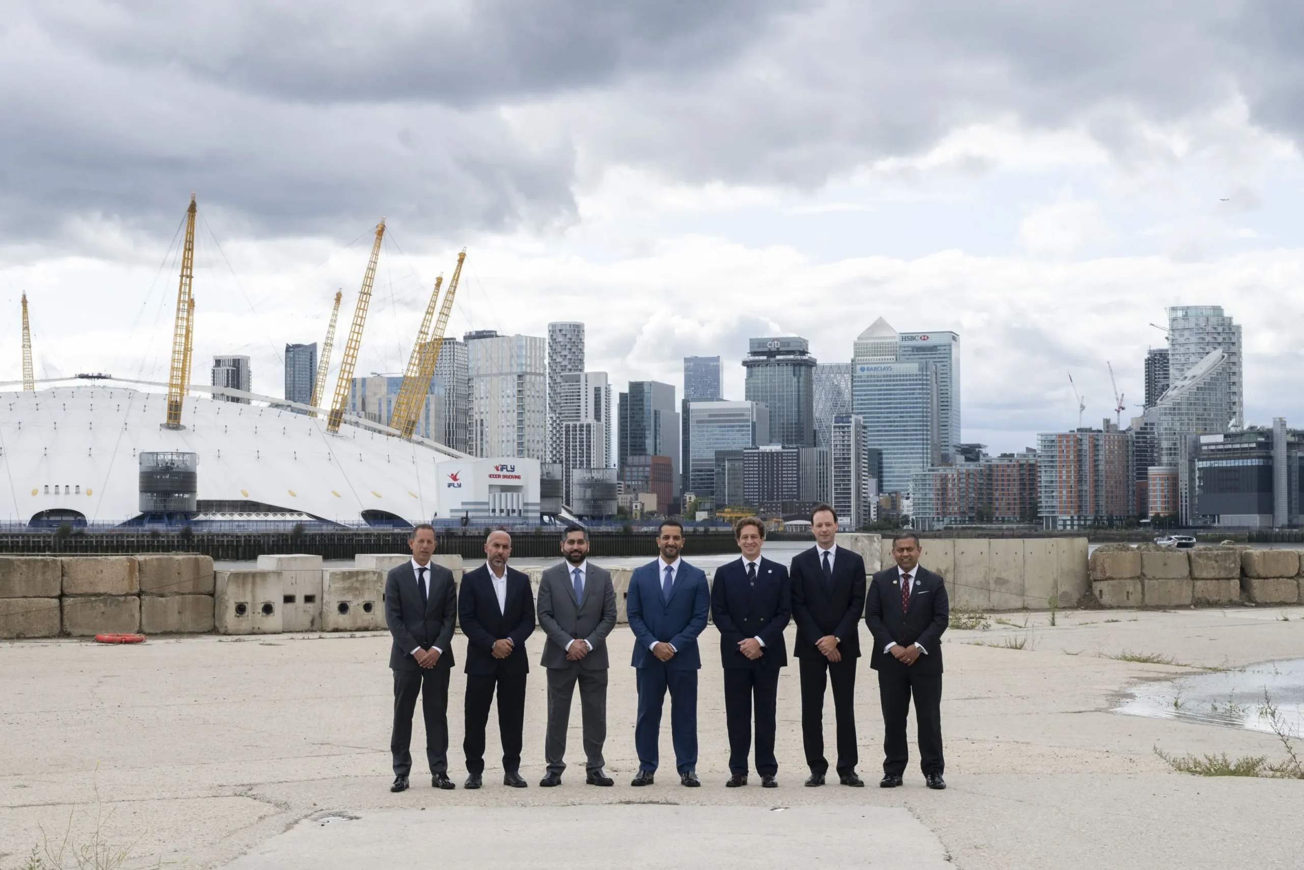 Seven men in business suits stand in a line on a concrete ground, facing the camera. Behind them is a modern city skyline with tall buildings, including the O2 Arena, on a cloudy day.
