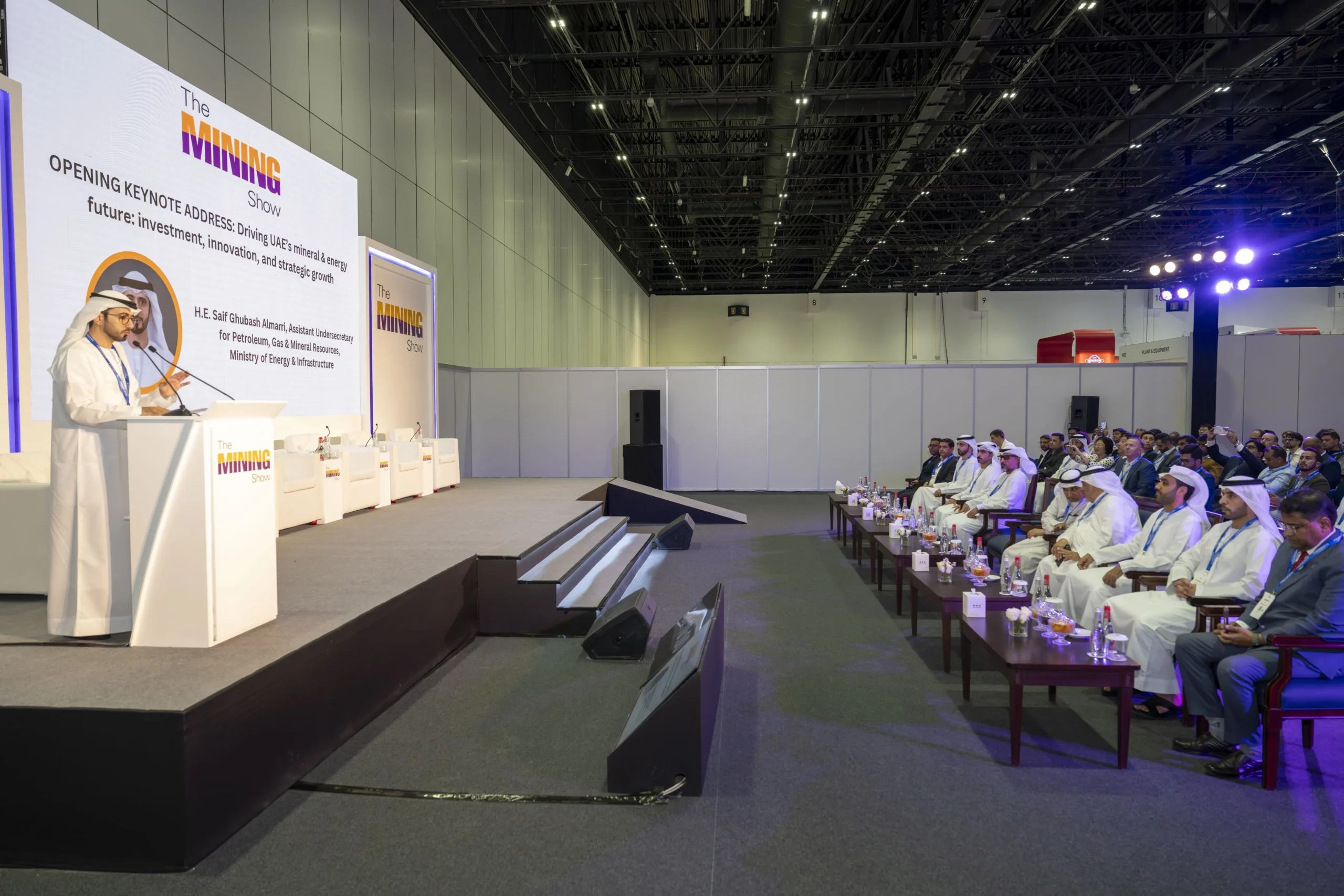 A man in traditional Emirati attire speaks at a podium with 'The Mining Show' logo on a large screen behind him, addressing an audience seated at tables in a large exhibition hall. The screen displays 'OPENING KEYNOTE ADDRESS: Driving UAE's mineral & energy future: investment, innovation, and strategic growth' and 'H.E. Saif Ghubash Almari, Assistant Undersecretary for Petroleum, Gas & Mineral Resources, Ministry of Energy & Infrastructure'.
