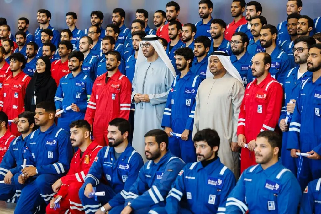 A large group of young men in blue and red work uniforms, some holding rolled diplomas, are gathered for a graduation ceremony. Two older men in traditional white Emirati thobes and ghutras stand prominently in the center, addressing the graduates. The setting appears to be an indoor event with a stage backdrop.