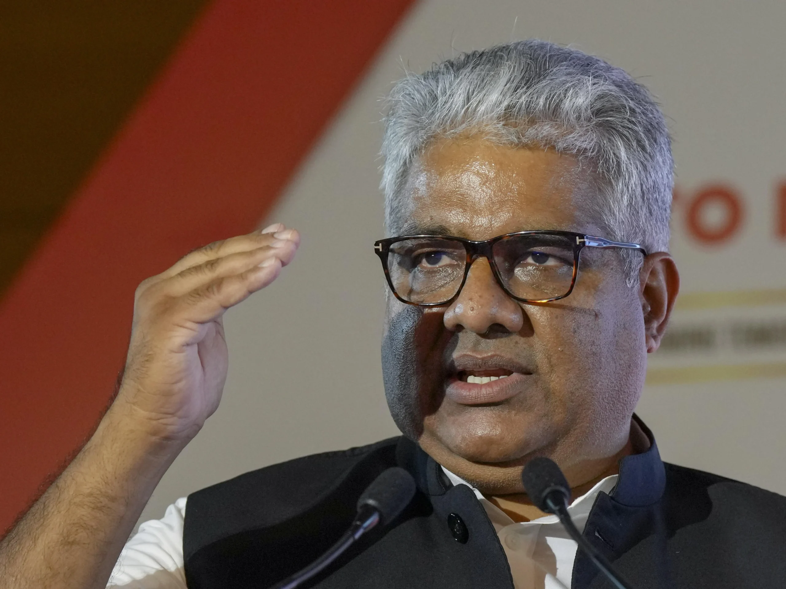 Union Environment Minister Bhupender Yadav, wearing glasses and a dark waistcoat over a white shirt, speaks animatedly into microphones at a podium, with his right hand raised in a gesturing motion. A red and white blurred background is visible behind him.