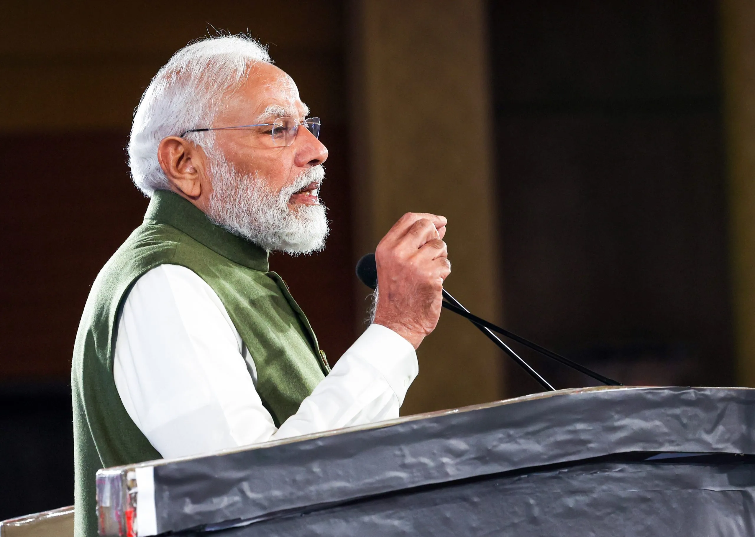Indian Prime Minister Narendra Modi speaks at a podium, holding a microphone in his right hand, wearing a white kurta and an olive green vest.