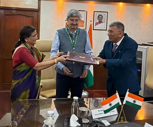 Dr. Samir V. Kamat, Chairman of DRDO, standing between Prof. Manoj Choudhary (Vice Chancellor, GSV) and Dr. Lal Chand Mangal (DS & DG(TM)), as a woman on the left and a man on the right exchange a signed document. The setting is an indoor office with flags of India on the table.