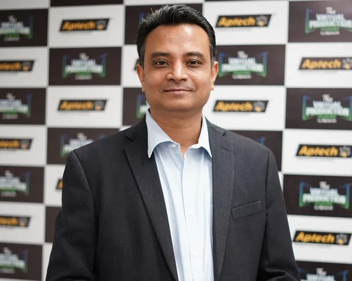 A man with short, dark hair, wearing a light blue collared shirt and a dark suit jacket, smiles slightly at the camera. He stands in front of a checkered black and white background with logos.