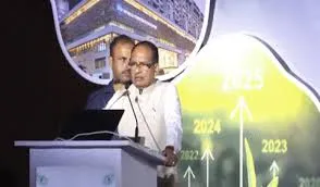Union Minister Shivraj Singh Chouhan speaking at a podium with a microphone, wearing a white kurta, addressing an event. A man in a blue shirt stands behind him. A projection screen in the background shows a building and a graph with years 2022, 2023, 2024, 2025.