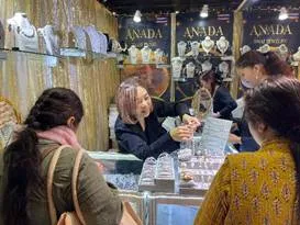 Ananda jewelry stall at a trade fair with women customers browsing necklaces and earrings. The stall features gold-colored backdrops and glass display cases filled with various jewelry pieces.