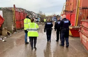Environment Agency officers and police personnel walk through a muddy, outdoor industrial site with large red waste containers and piles of scrap metal and waste in the background.
