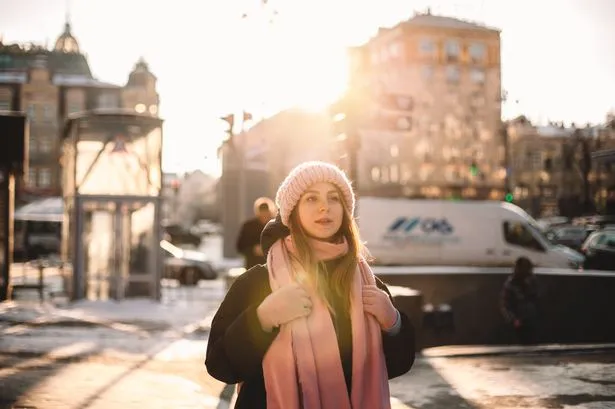 A young woman wearing a pink knitted hat, a long pink scarf, and a dark coat stands outdoors in what appears to be a city setting during winter. The sun is bright, creating a strong backlight and lens flare, illuminating her hair and the edges of her scarf. Buildings and some vehicles are visible in the blurred background. She is looking slightly to her right with a neutral expression.