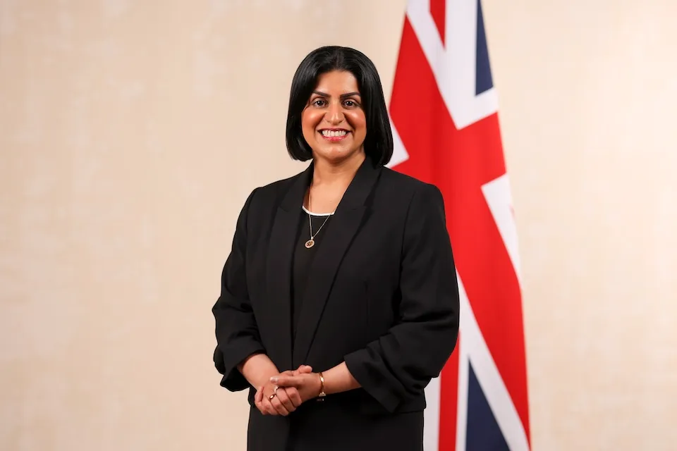 Shabana Mahmood MP, dressed in a black blazer over a dark top, stands smiling with her hands clasped in front of a light cream wall and a partially visible Union Jack flag.