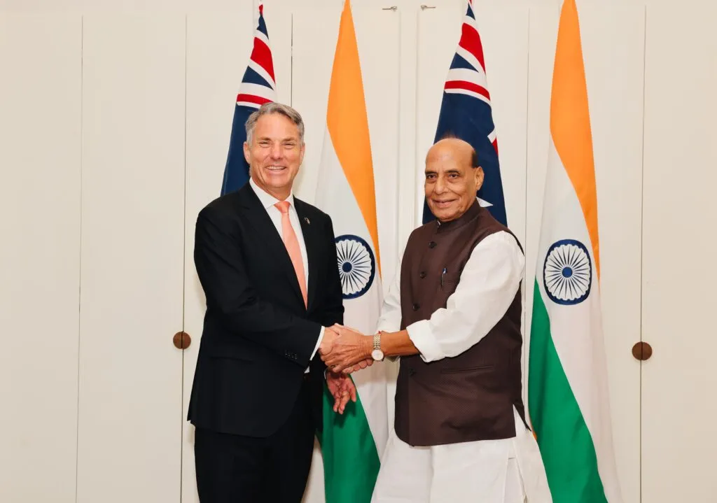 Australian Defence Minister Richard Marles and Indian Defence Minister Rajnath Singh shake hands in front of Australian and Indian flags.
