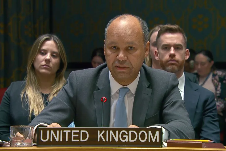 James Kariuki, UK Chargé d’Affaires to the UN, is seated at a table with a 'UNITED KINGDOM' nameplate in front of him, speaking at a UN Security Council meeting. He is wearing a dark suit and a light blue tie. Several other people are visible blurred in the background.