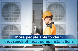A man in an orange high-visibility jacket and a yellow hard hat is standing in front of large outdoor air conditioning units or heat pumps. He is holding a tablet or clipboard and appears to be inspecting or working on the equipment. The text overlay on the image reads 'More people able to claim thousands off heat pump installations'.