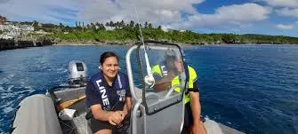 Two individuals in a small boat on clear blue water, with one person in the foreground facing the camera and wearing a life vest. Green foliage and buildings are visible on a distant shoreline under a partly cloudy sky.