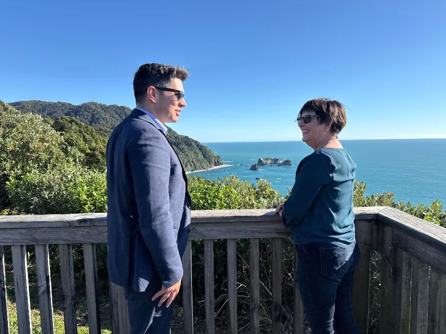 New Zealand Associate Transport Minister James Meager and local MP Maureen Pugh standing on a wooden observation deck overlooking the Tasman Sea and rugged coastline near the Epitaph Slip site on State Highway 6. The sky is clear and blue, and the ocean extends to the horizon with notable rock formations. Lush green vegetation covers the foreground and cliffs.