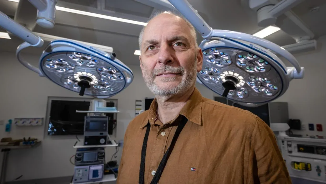 A man with a short beard and mustache, wearing a brown collared shirt, stands in what appears to be a modern operating room. Two large, round surgical lights with multiple LED bulbs hang from the ceiling behind him. Various medical equipment, including monitors and carts, are visible in the background, out of focus.